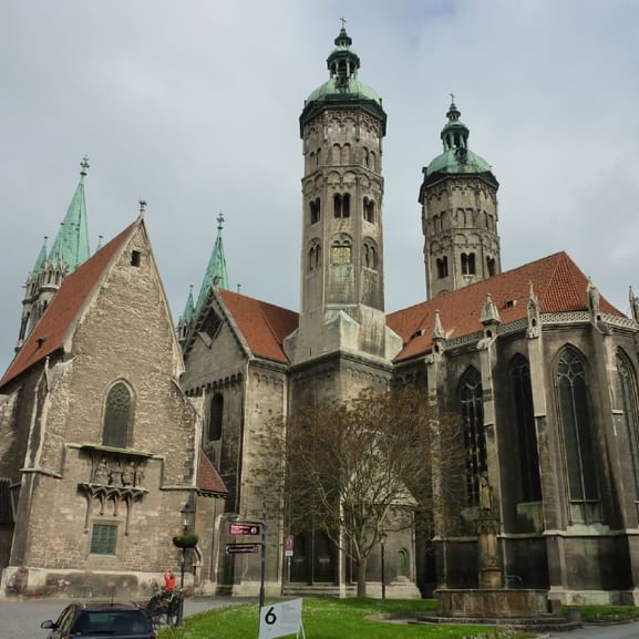 Naumburger Dom mit Doppeltürmen Naumburger Dom mit zwei markanten Steintürmen mit grünen Kupferkuppeln, roten Ziegeldächern und gotischer Architektur vor bewölktem Himmel.