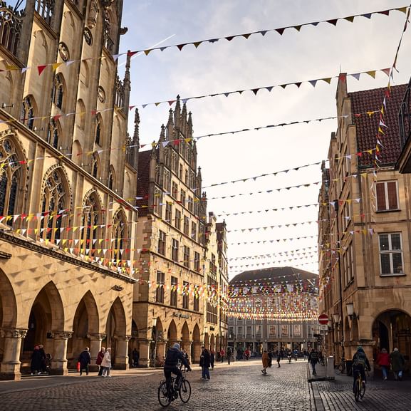 Historischer Prinzipalmarkt in Münster mit gotischem Rathaus, Arkadenhäusern und bunten Wimpeln. Radfahrer und Fußgänger überqueren den Kopfsteinplatz.