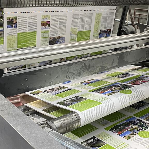 Industrielle Druckmaschine produziert farbige Kataloge mit grünen kreisförmigen Elementen und Reisebildern auf weißen Papierbögen.