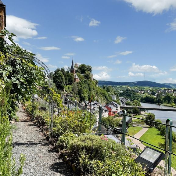 Schmaler Kiesweg durch Saarburg mit Weinbergterrassen, Metallgeländer, Blick auf die Saar und Stadt mit Kirchturm.