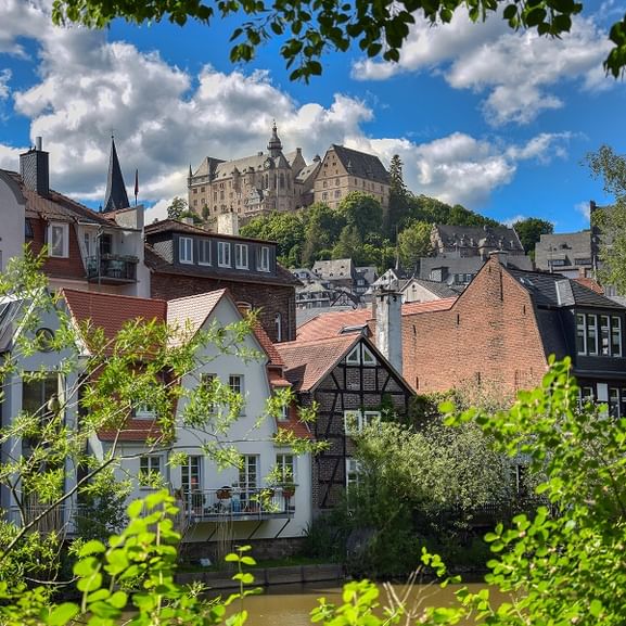 View of Marburg's historic town center with traditional half-timbered houses along the river, dominated by the medieval castle on the hill above.