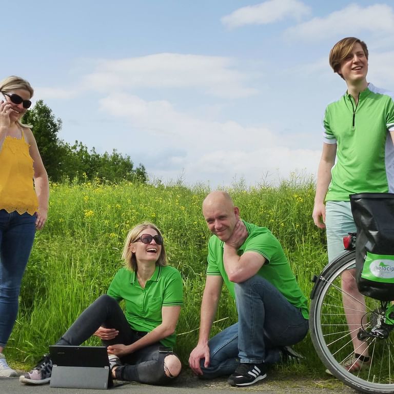 Vier Velociped Teammitglieder auf grüner Wiese. Frau im gelben Top telefoniert, zwei Personen in grünen Shirts sitzen, Mann mit Fahrrad.